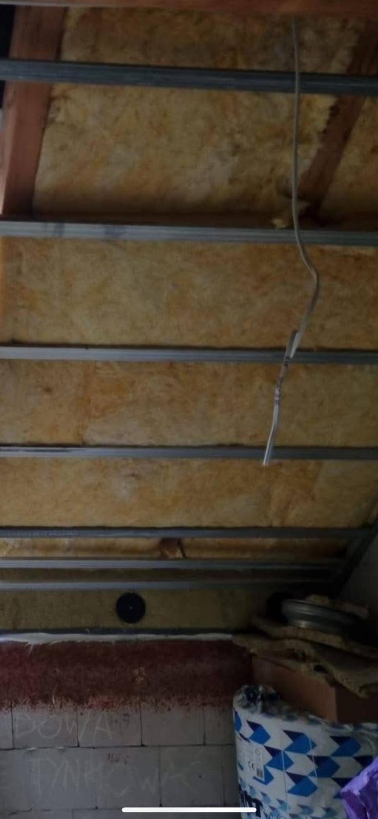 Montaż izolacji z wełny mineralnej w poddaszu, widoczne profile metalowe konstrukcji sufitu podwieszanego i wystający kabel elektryczny.