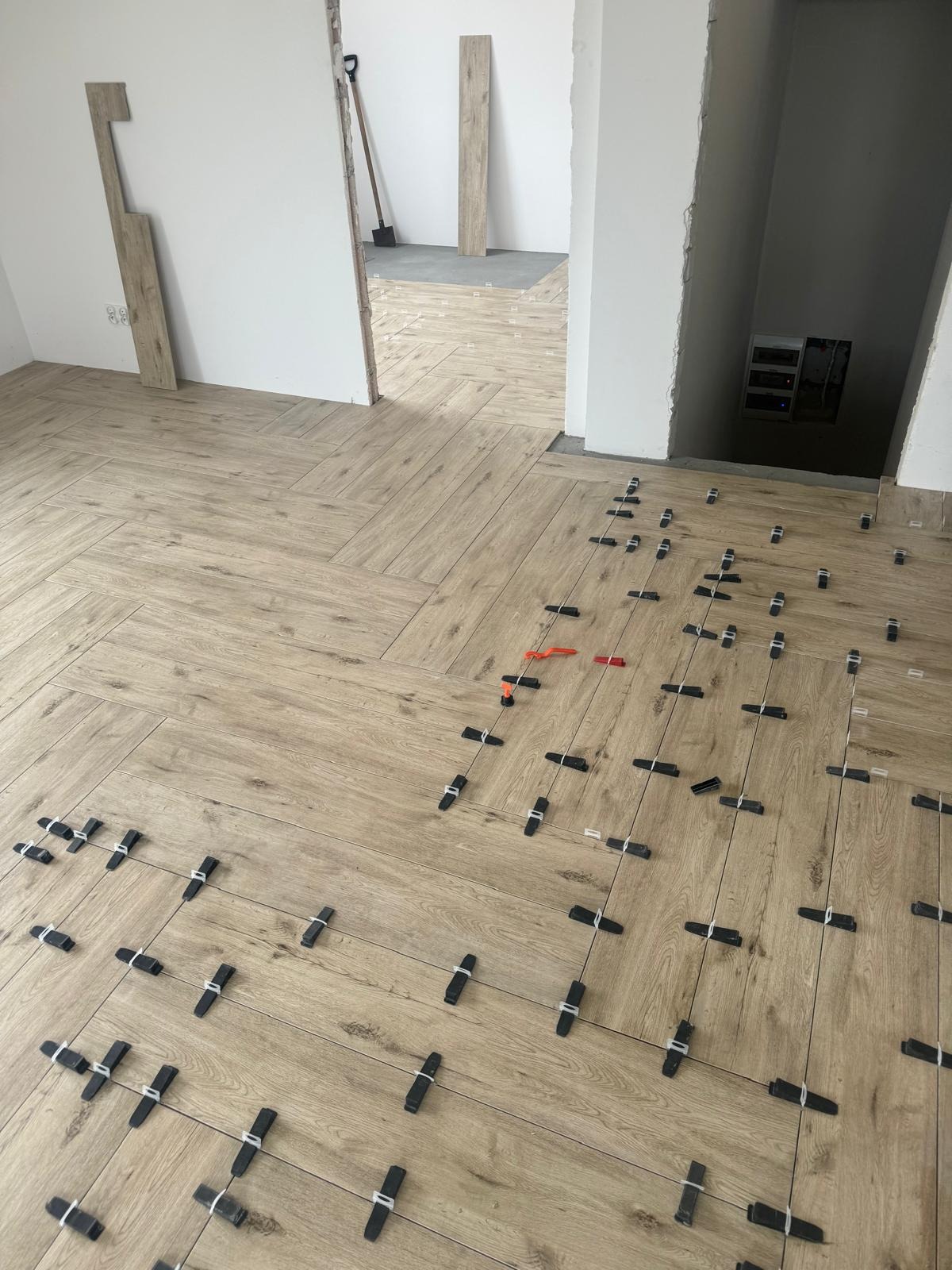 Układanie płytek podłogowych imitujących drewno w jodełkę, z widocznymi klipsami poziomującymi i sznurkami dystansowymi, fragment ściany z niedokończonymi drzwiami i narzędzia w tle.