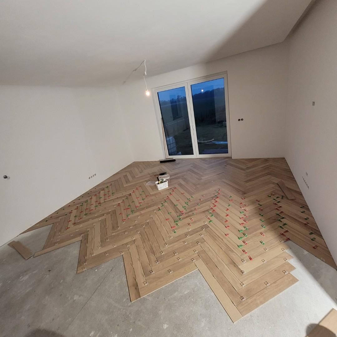 Układanie parkietu w jodełkę, widoczne białe ściany, okno i kliny dystansowe w kolorach czerwonym i zielonym.