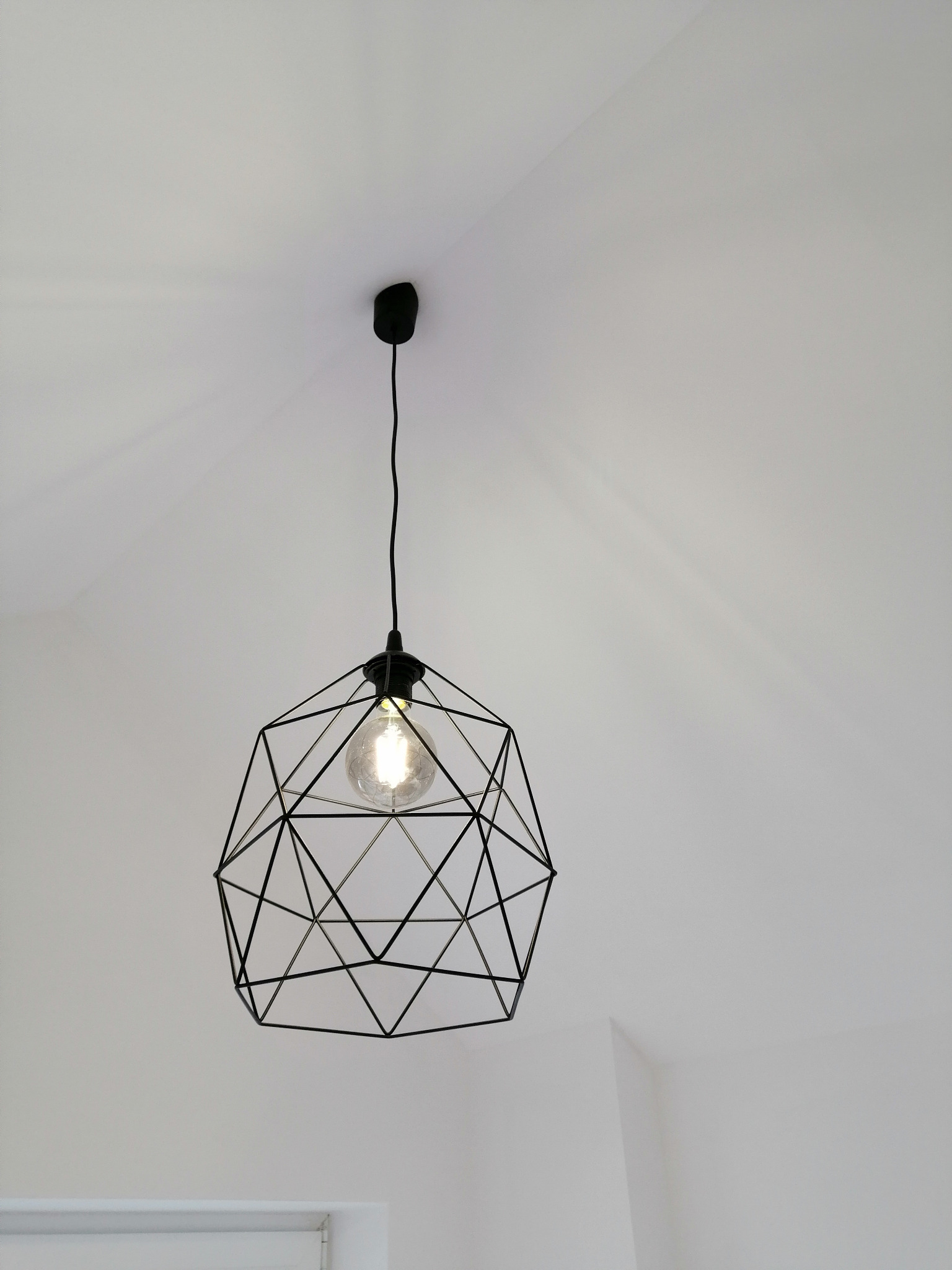 Czarna, geometryczna lampa wisząca z żarówką Edison w minimalistycznym wnętrzu. Kąt ujęcia od dołu podkreśla nowoczesny design lampy.