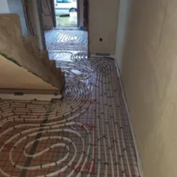 Rozłożone rury ogrzewania podłogowego na folii z siatką, widok z góry na etap instalacji w surowym wnętrzu domu.