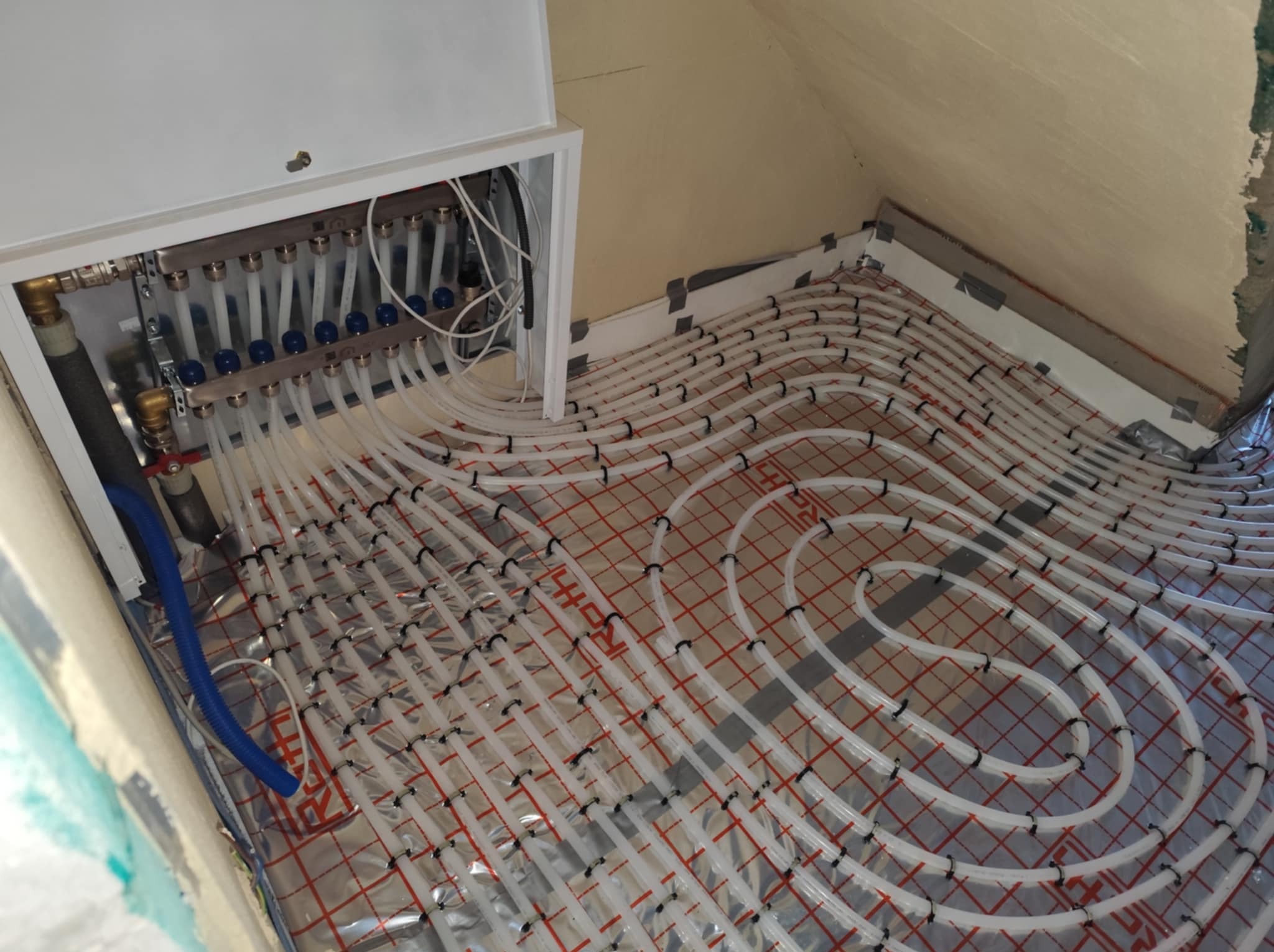 Rozłożona instalacja ogrzewania podłogowego w trakcie montażu, widoczne białe rurki grzewcze zamocowane na folii z siatką, rozdzielacz z zaworami w białej obudowie na ścianie, surowy stan ścian.