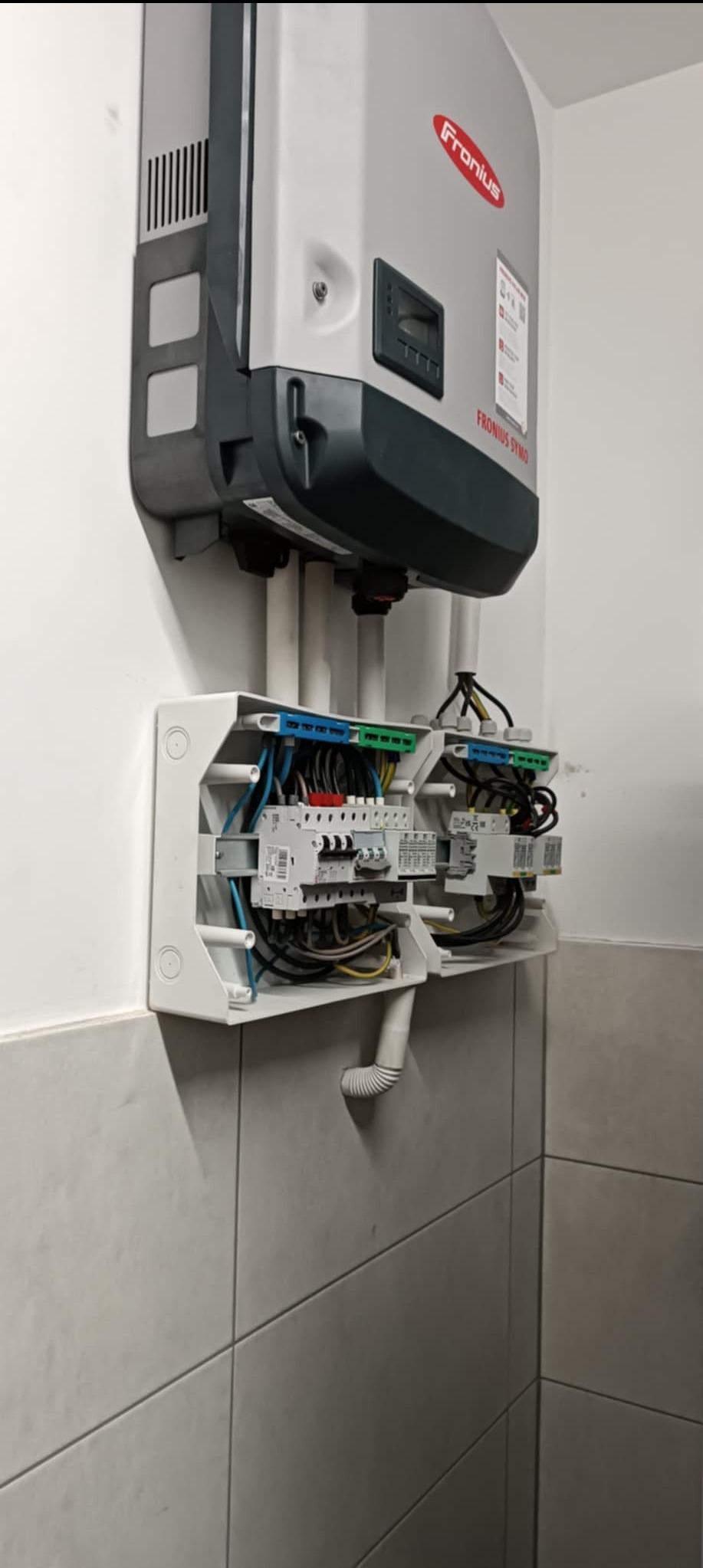 Szary inwerter Fronius SYMO zamontowany na białej ścianie nad otwartą skrzynką z bezpiecznikami i okablowaniem w kolorach niebieskim, zielonym, żółtym i czarnym. Widoczne rurki PCV prowadzące kable.