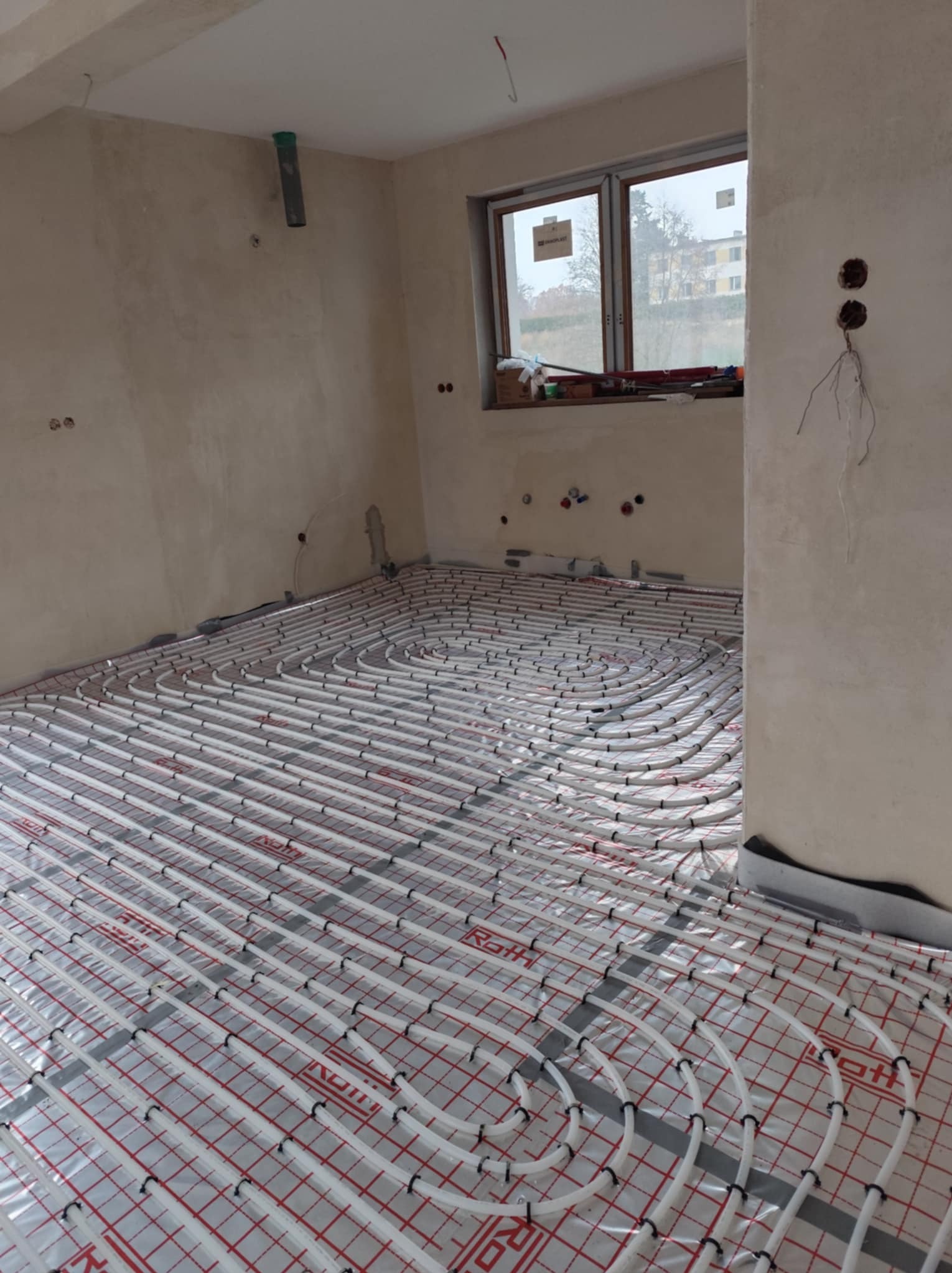 Rozłożone rury ogrzewania podłogowego marki Roth na folii z czerwoną kratką w surowym pomieszczeniu z widocznymi puszkami elektrycznymi i oknem.