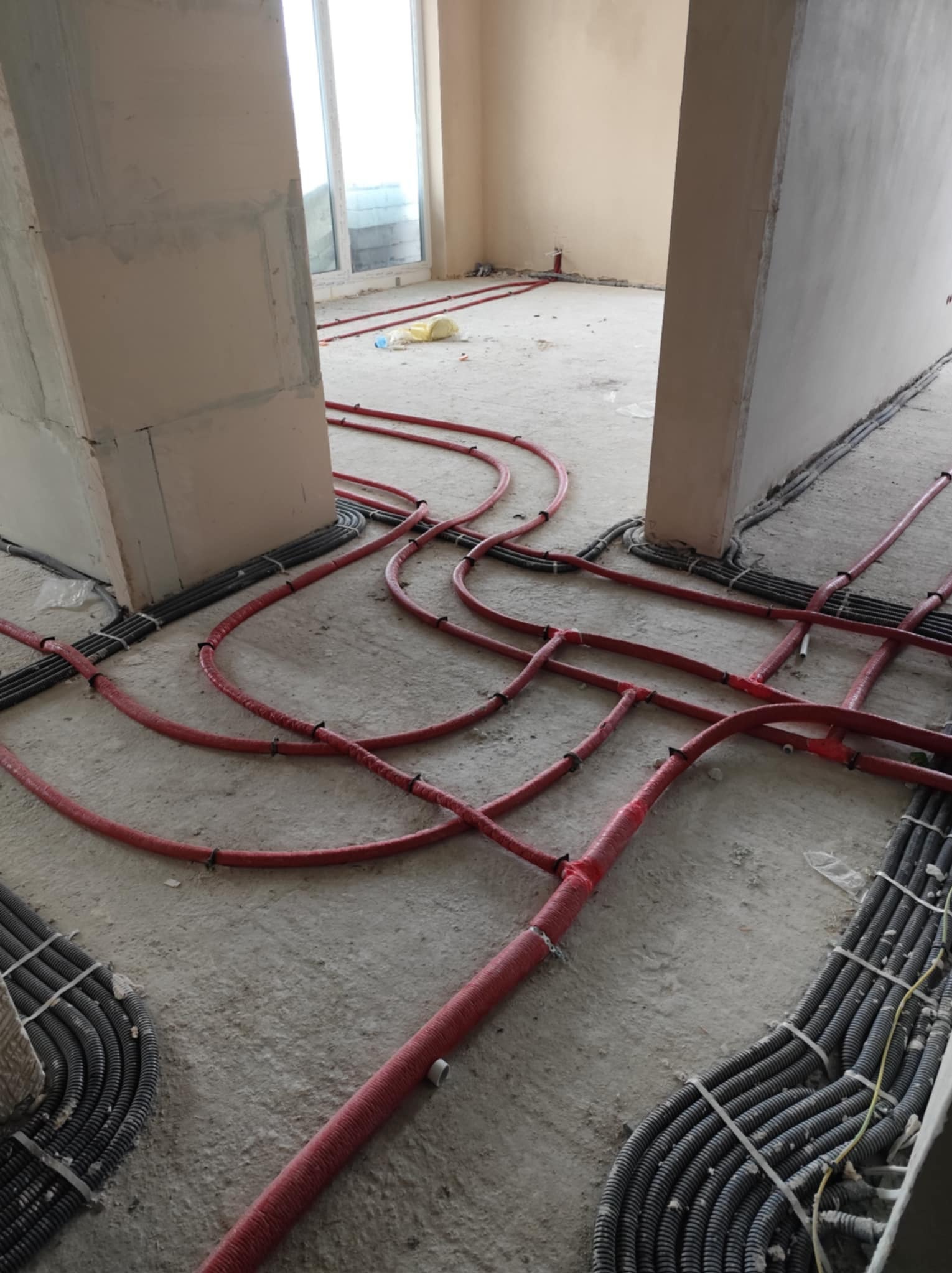 Instalacja ogrzewania podłogowego w wylewce, widoczne czerwone rury grzewcze i czarne rury osłonowe kabli elektrycznych, surowy stan wykończenia.