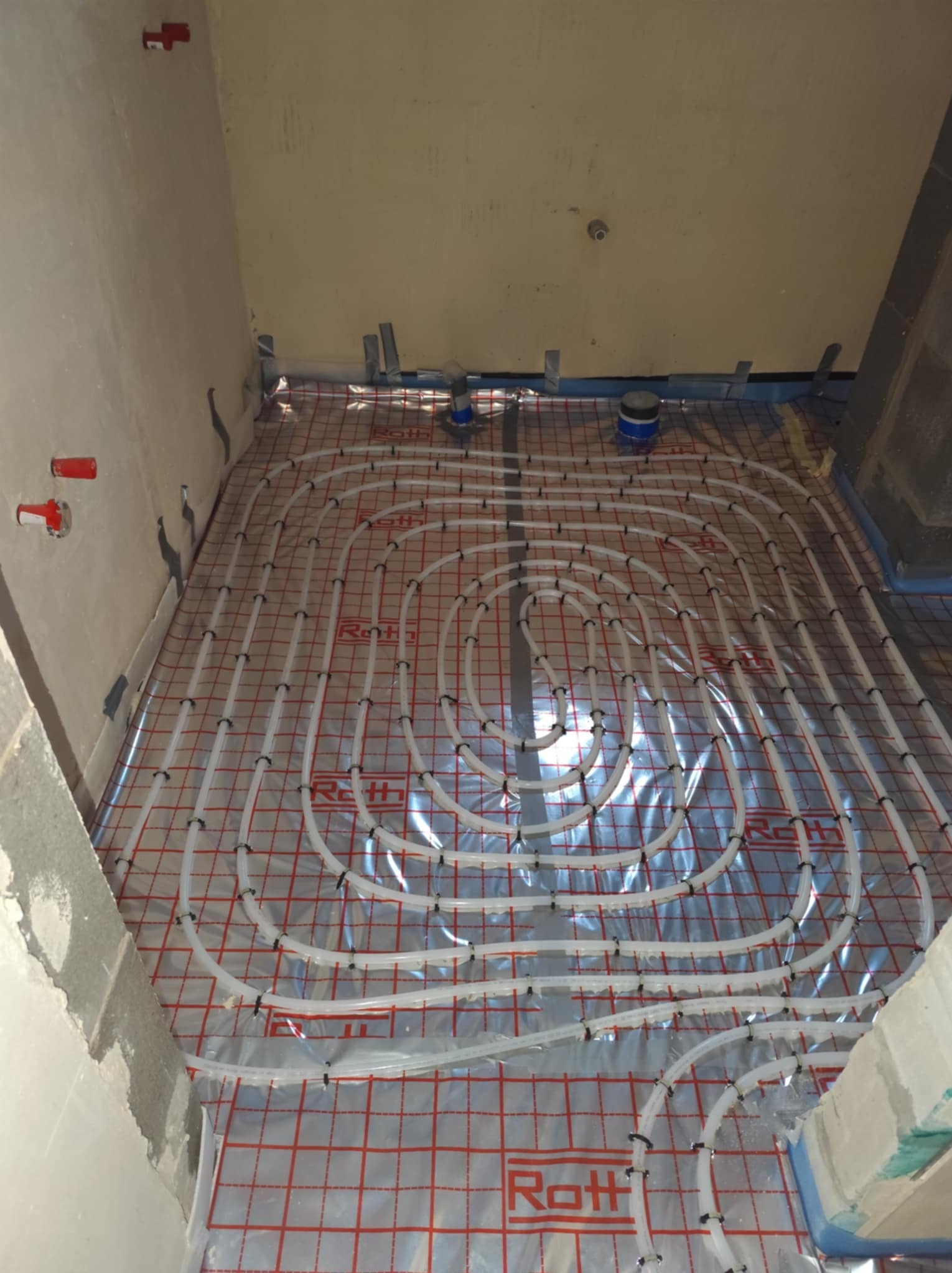 Białe rury ogrzewania podłogowego ułożone spiralnie na folii z czerwoną siatką w surowym, przygotowywanym do wykończenia pomieszczeniu z widocznymi elementami instalacji na ścianach.