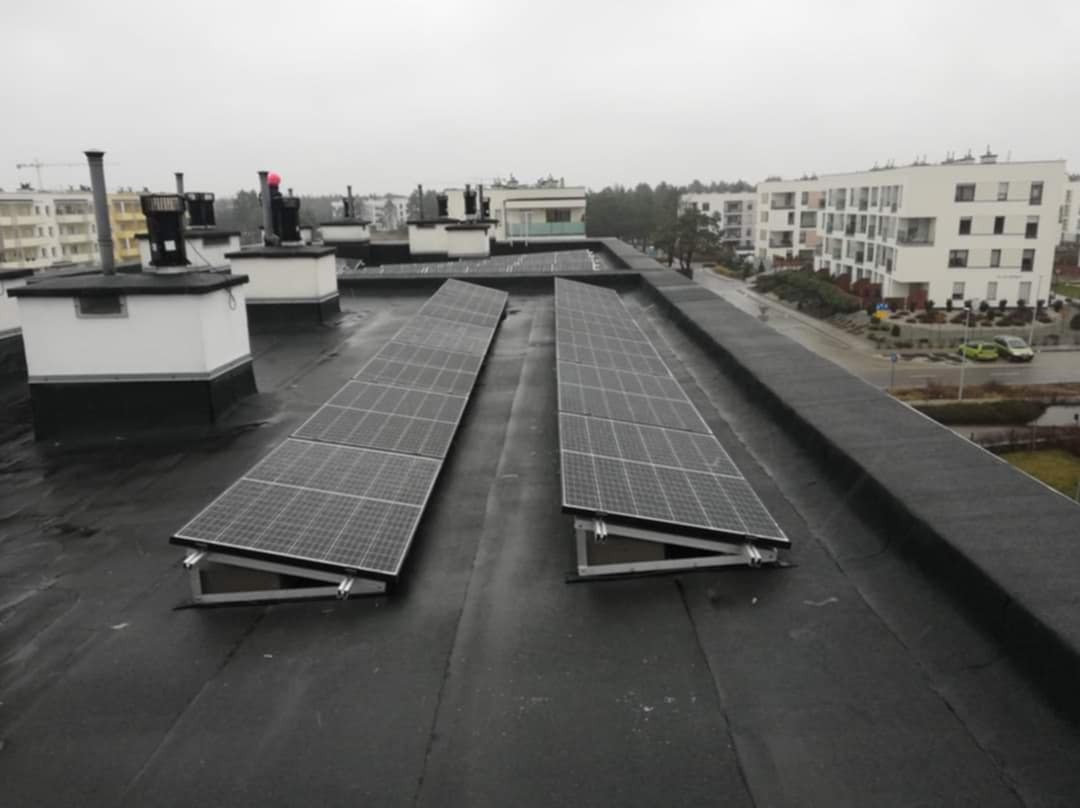 Dwa rzędy paneli słonecznych zamontowane na płaskim, czarnym dachu budynku mieszkalnego w pochmurny dzień. Widoczne konstrukcje wsporcze, kominy wentylacyjne i zabudowa osiedlowa w tle.