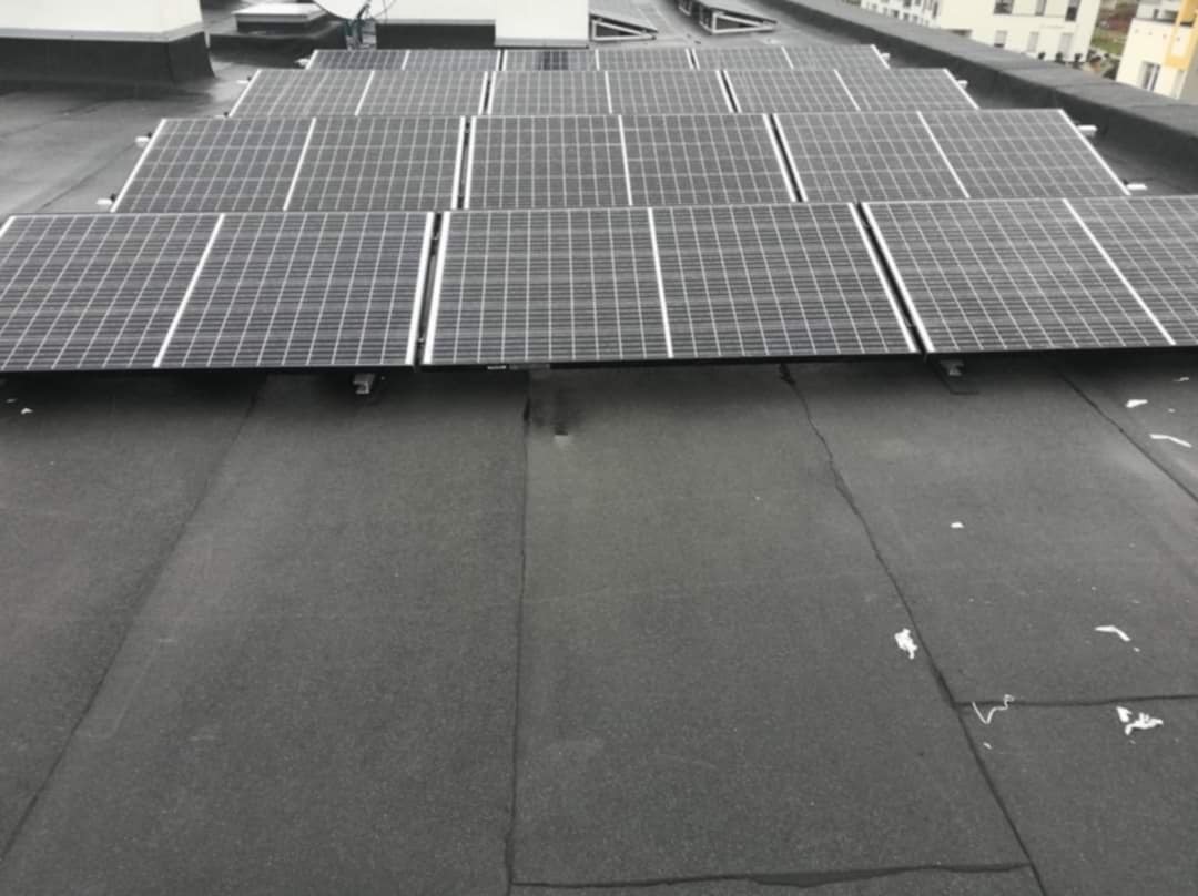 Rząd paneli słonecznych zamontowanych na płaskim, bitumicznym dachu budynku mieszkalnego, widoczne z góry pod lekkim kątem. Na dachu widoczne drobne zabrudzenia i łączenia papy.
