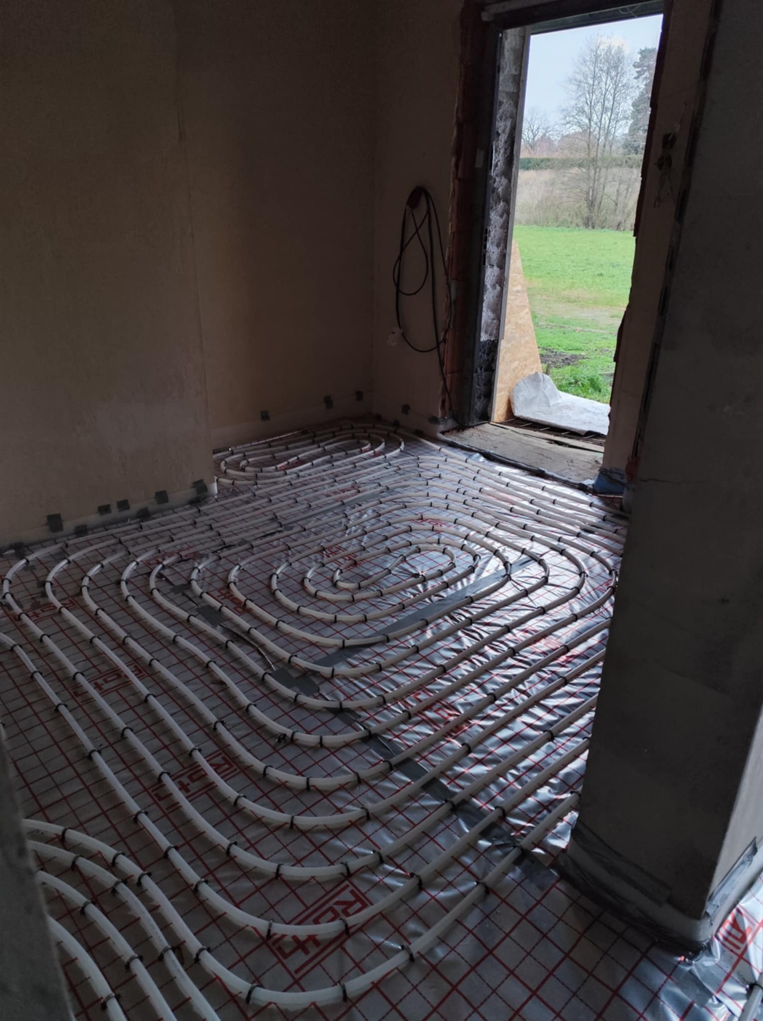 Rozłożona instalacja ogrzewania podłogowego z białych rurek na folii z czerwoną siatką w surowym pomieszczeniu z widokiem na zewnątrz przez otwarte drzwi balkonowe.