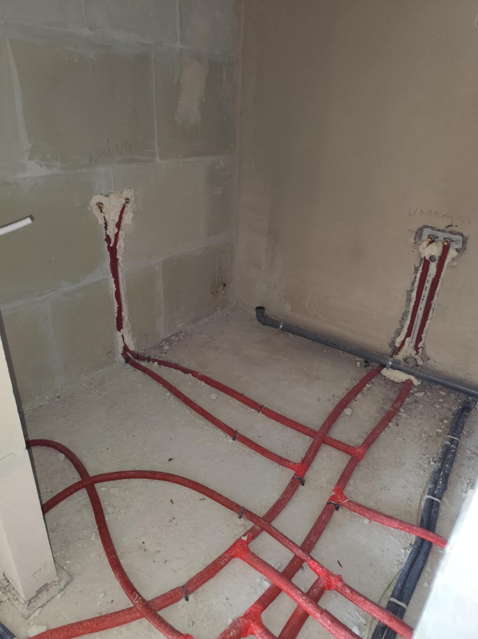 Wnętrze w trakcie prac hydraulicznych: czerwone rury grzewcze podłogowe ułożone na surowym betonie, pionowe instalacje w ścianach wypełnione pianką montażową, widoczne napisy 'Wanna' i 'Umywalka'...