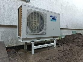 Zewnętrzna jednostka pompy ciepła marki Gree zamontowana na metalowej konstrukcji przy ścianie budynku, widoczna ziemia i pozostałości po pracach instalacyjnych.