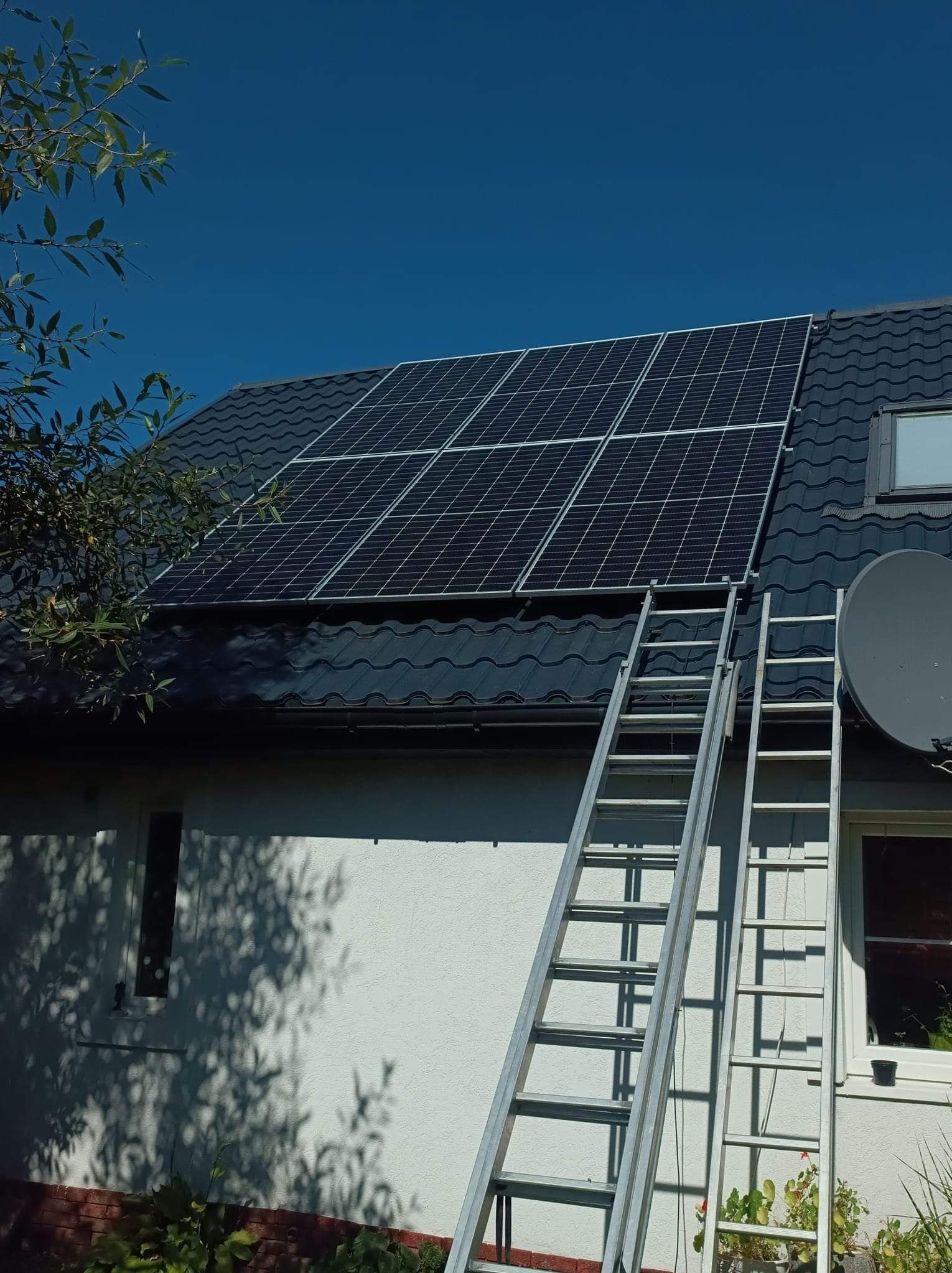 Panele fotowoltaiczne zamontowane na dachu domu jednorodzinnego z ciemną dachówką, widoczne dwie aluminiowe drabiny opierające się o ścianę budynku, błękitne niebo w tle.