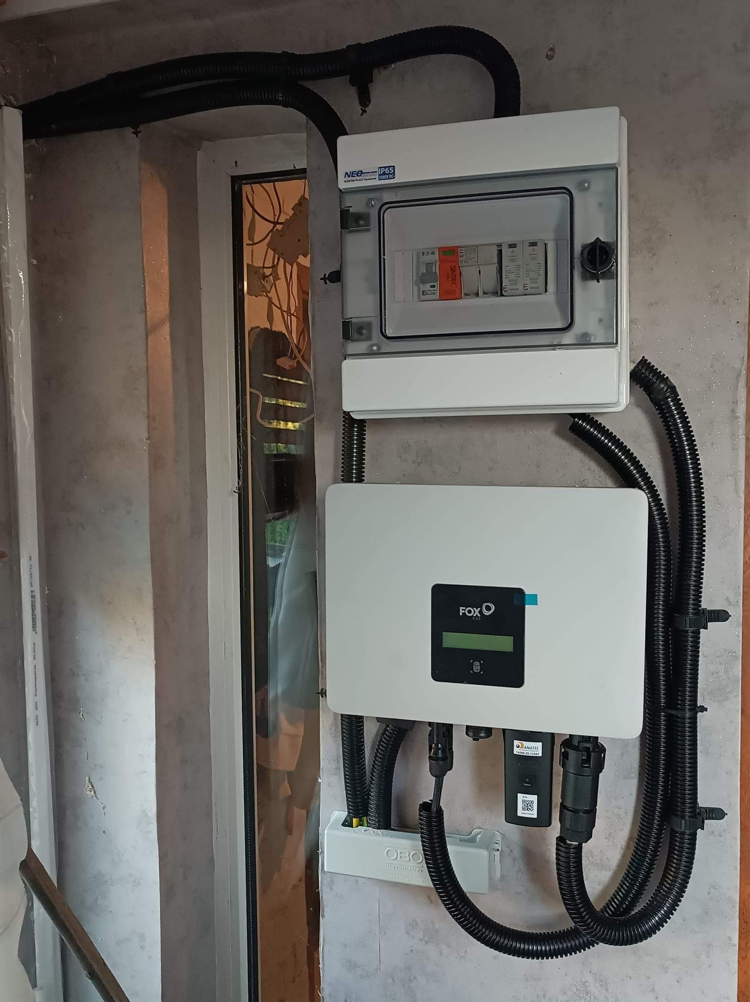 Panel fotowoltaiczny FOX i skrzynka z bezpiecznikami zamontowane na ścianie w Toruniu, widoczne czarne rury kablowe i okno.