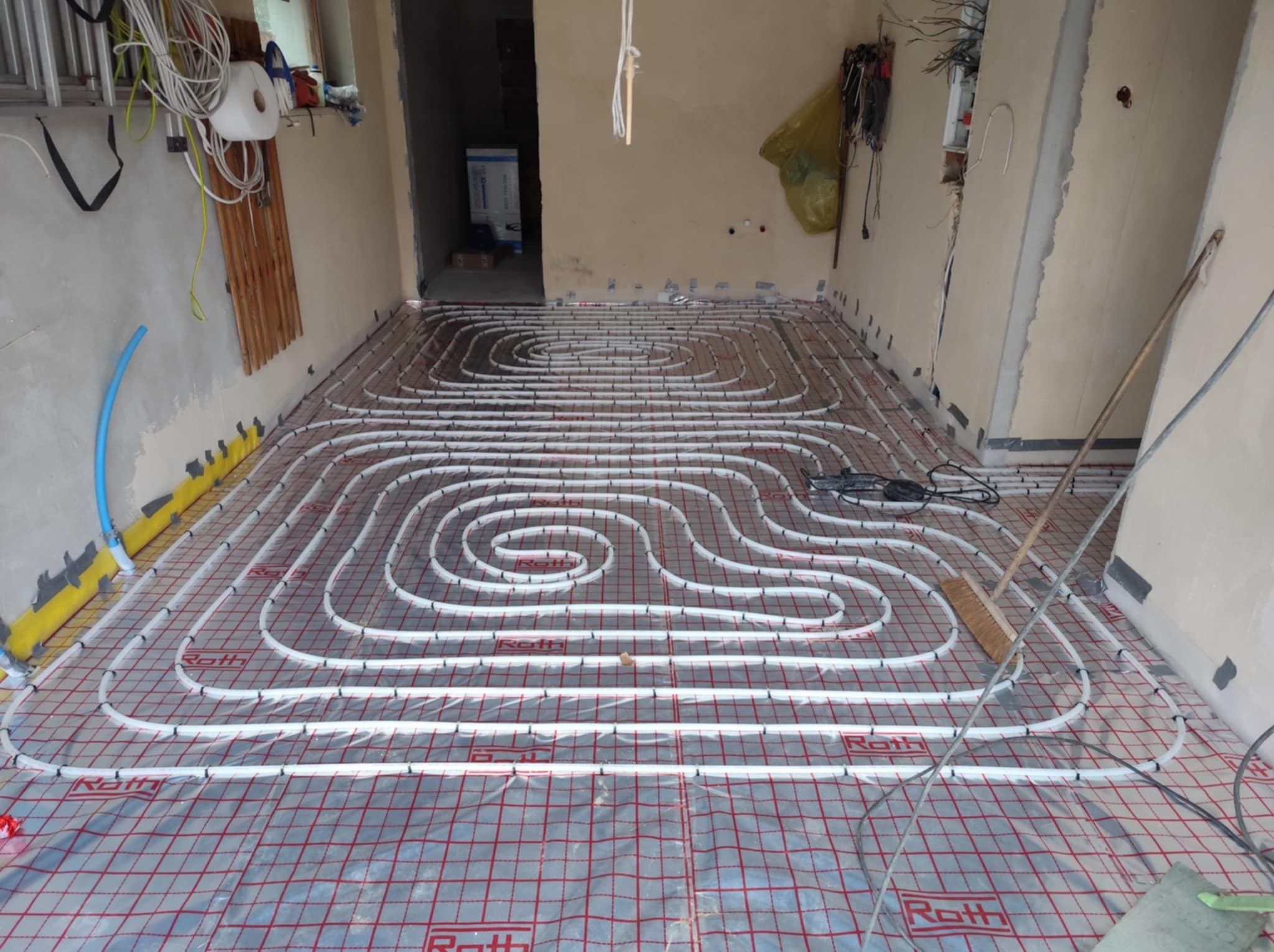 Białe rury systemu ogrzewania podłogowego Roth ułożone spiralnie na folii z czerwoną siatką w surowym wnętrzu przed wylewką, widoczne narzędzia i kable elektryczne.