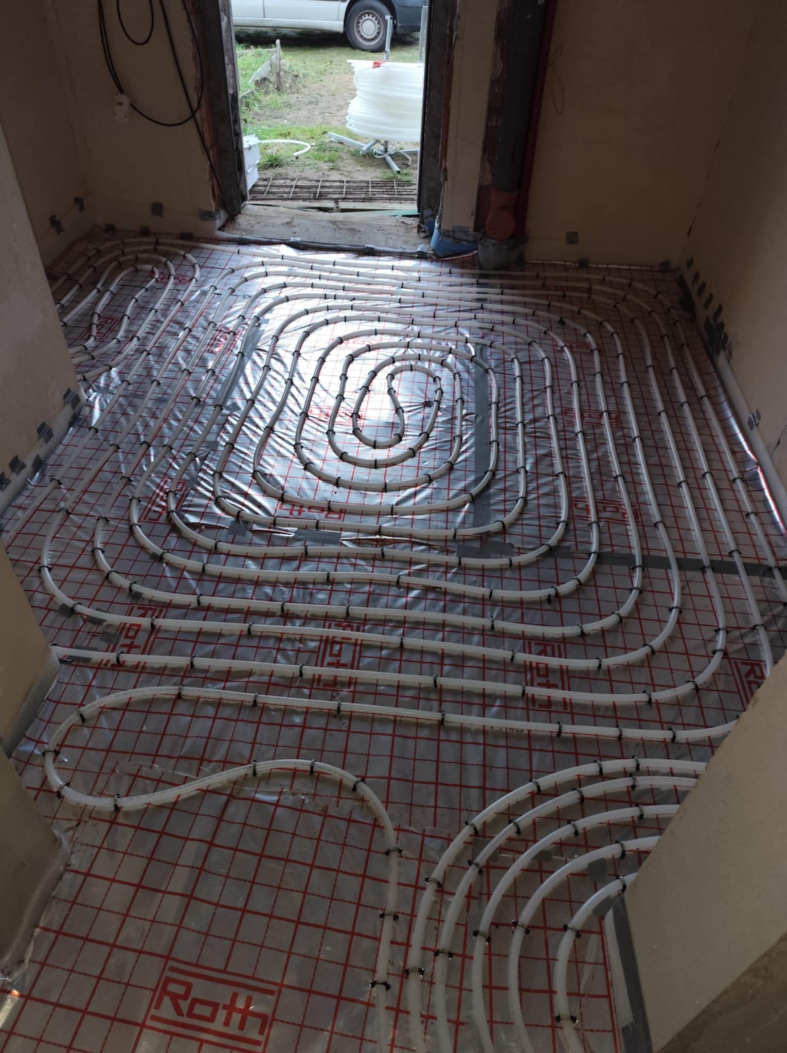 Rozłożone białe rury ogrzewania podłogowego na folii z czerwoną kratką, widoczne z góry w surowym wnętrzu podczas instalacji, marka Roth widoczna w dolnym rogu.