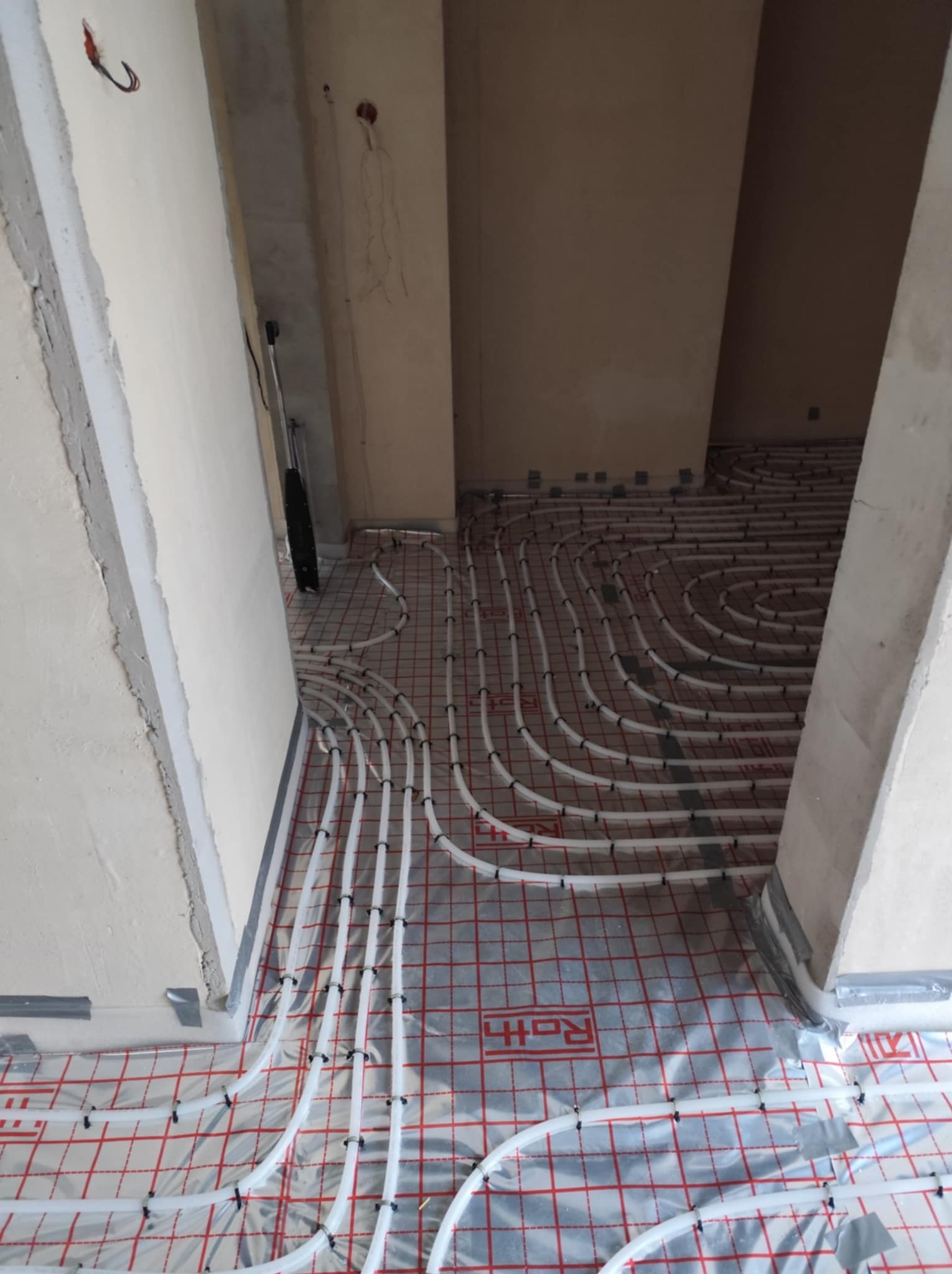 Rozłożone białe rury ogrzewania podłogowego na folii z czerwoną kratką, widok z góry na surowy stan wykończenia wnętrza.