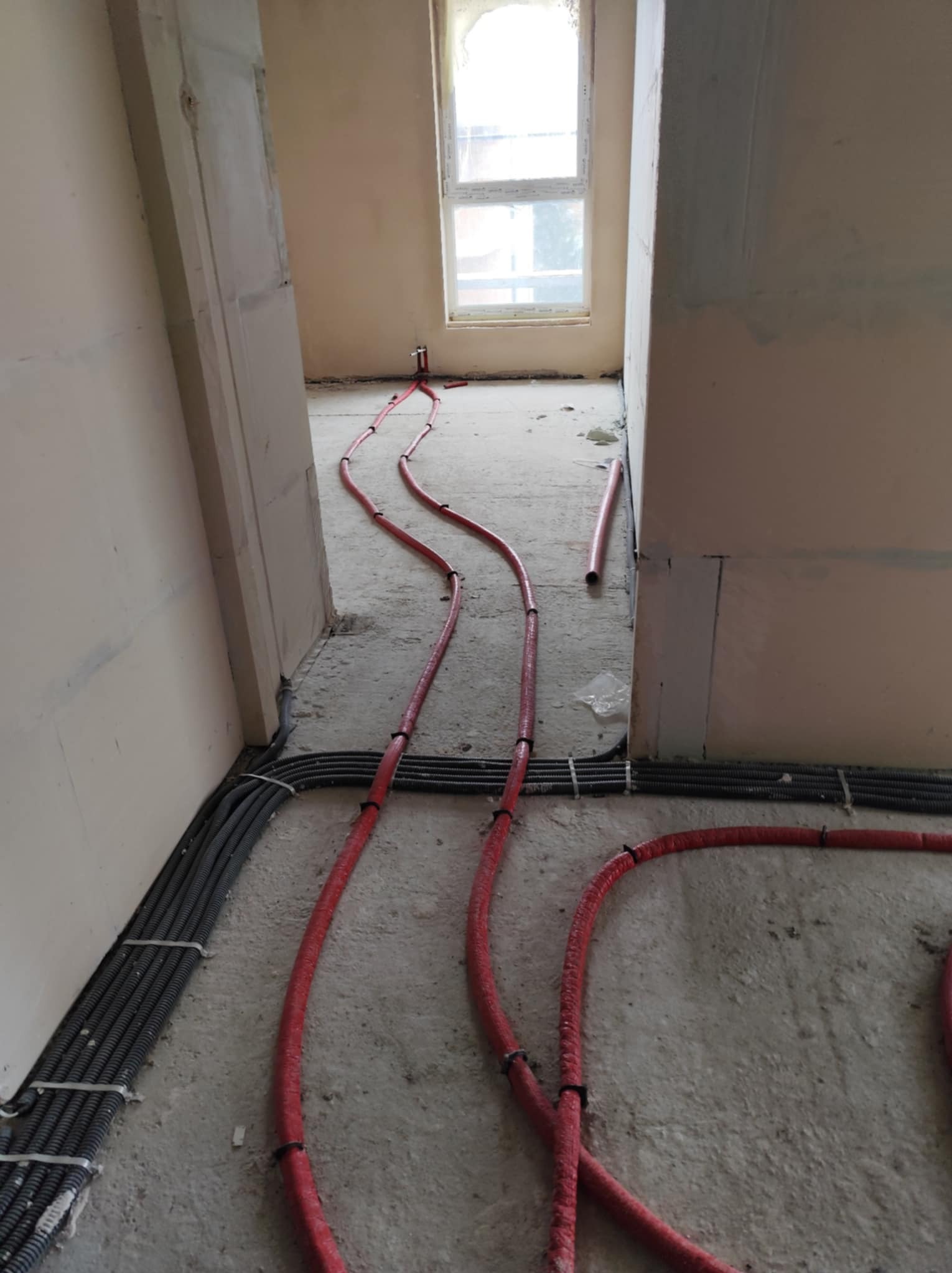 Wnętrze w trakcie prac wykończeniowych z widocznymi czerwonymi rurami ogrzewania podłogowego ułożonymi na szarym, surowym betonie oraz szarymi przewodami elektrycznymi przy ścianach.