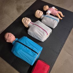 Safe Work Maciej Łusiak - Manekiny do nauki pierwszej pomocy, w tym niemowlę, leżą na czarnej macie obok czerwonej torby. Widok z góry, oświetlenie studyjne.