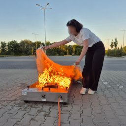 Safe Work Maciej Łusiak - Osoba demonstruje gaszenie płonącego materiału na metalowej wannie podczas szkolenia ppoż. Widoczne płomienie, materiał i wąż doprowadzający paliwo.
