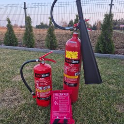 Safe Work Maciej Łusiak - Dwie gaśnice (proszkowa i CO2) oraz koc gaśniczy na trawie, na tle ogrodzenia i tuj. Sprzęt ppoż. w otoczeniu zewnętrznym.