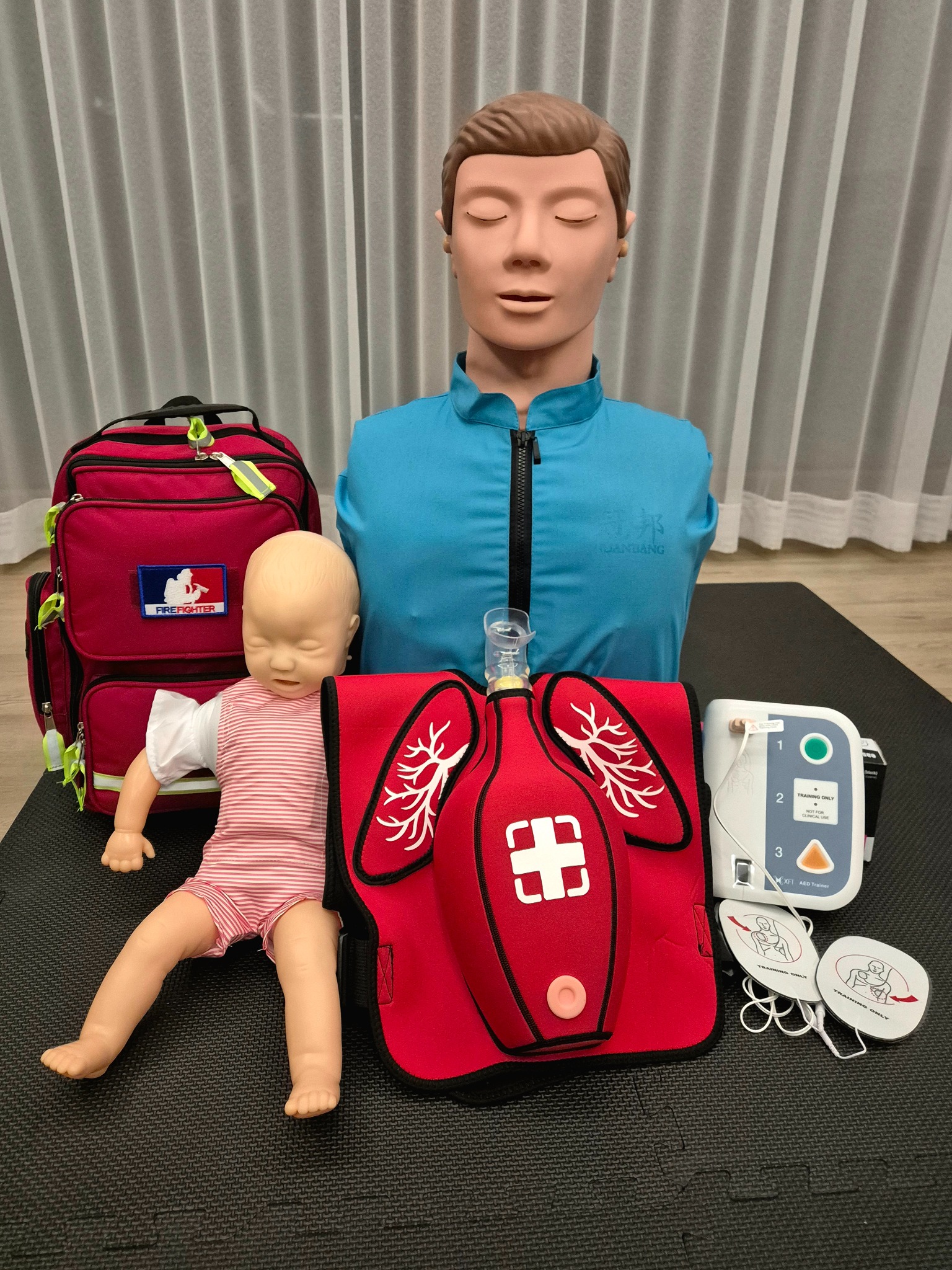 Manekin do nauki RKO, fantom niemowlęcy, defibrylator treningowy i torba ratownicza na czarnej macie. Sprzęt do kursu pierwszej pomocy.