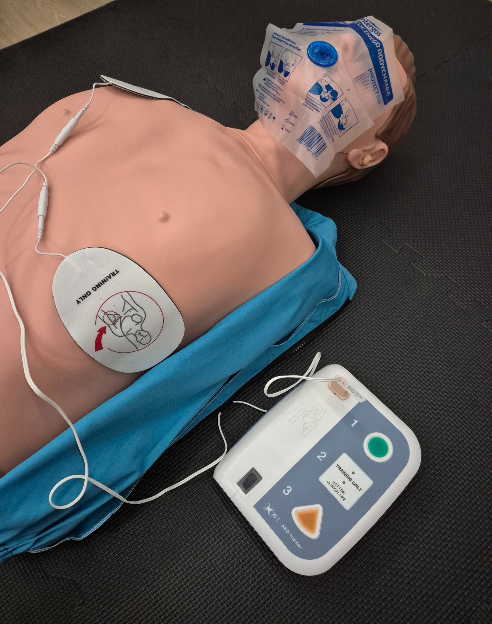 Manekin do nauki pierwszej pomocy z podłączonym defibrylatorem treningowym i maską twarzową. Sprzęt do ćwiczeń resuscytacji krążeniowo-oddechowej.