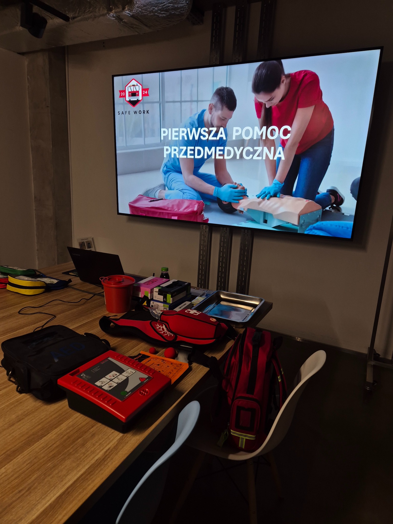 Sala szkoleniowa z ekranem prezentującym kurs pierwszej pomocy. Na stole torby medyczne, AED, manekin. Widoczny laptop i krzesło. Szkolenie z zakresu ratownictwa.
