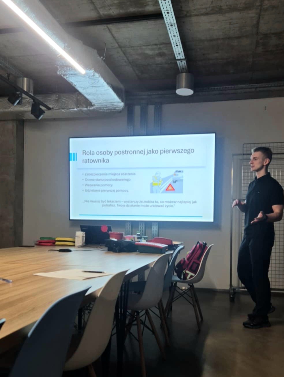Prelegent omawia rolę osoby postronnej jako ratownika podczas kursu pierwszej pomocy. Na ekranie slajd z zasadami udzielania pomocy. Sala konferencyjna.