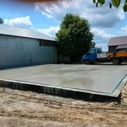 Hulewscy Spółka Cywilna - Świeżo wylana, gładka płyta betonowa fundamentowa z widocznymi deskami szalunkowymi na obrzeżach, na piaszczystym podłożu, z budynkami gospodarczymi w tle i błękitnym niebem z chmurami.