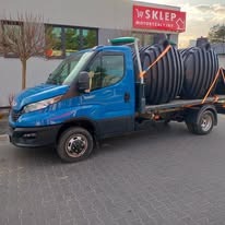 Niebieski samochód dostawczy Iveco z trzema czarnymi zbiornikami z tworzywa sztucznego, zabezpieczonymi pomarańczowymi pasami transportowymi, zaparkowany na kostce brukowej przed budynkiem sklepu.