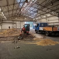 Mini koparka pracująca wewnątrz hali, usuwająca ziemię z pryzmy, obok stoi mała ciężarówka z otwartą skrzynią ładunkową, w tle widoczna konstrukcja dachu hali.