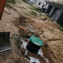 Wykonany wykop pod zbiornik z zieloną pokrywą, widoczne rury kanalizacyjne i fragment łyżki koparki, teren budowy.