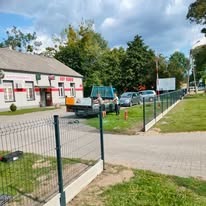 Montaż ogrodzenia panelowego na podmurówce prefabrykowanej, widoczny samochód ciężarowy z wywrotką oraz budynek w tle.