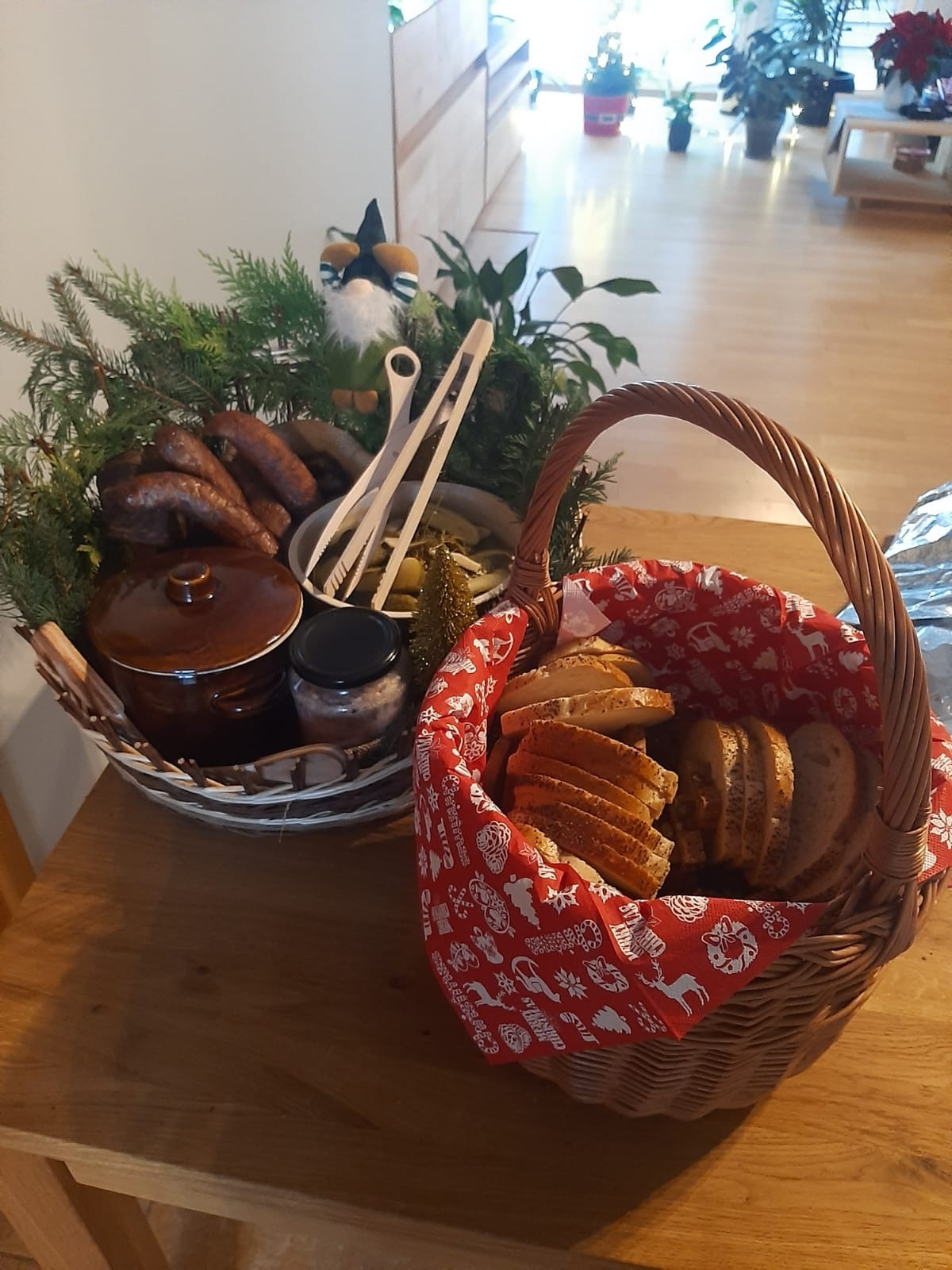Dwa wiklinowe kosze z jedzeniem: jeden z pieczywem w czerwonej serwetce z motywami świątecznymi, drugi z wędlinami, garnkiem, słoikiem, ogórkami kiszonymi, szczypcami i dekoracjami z gałązek...