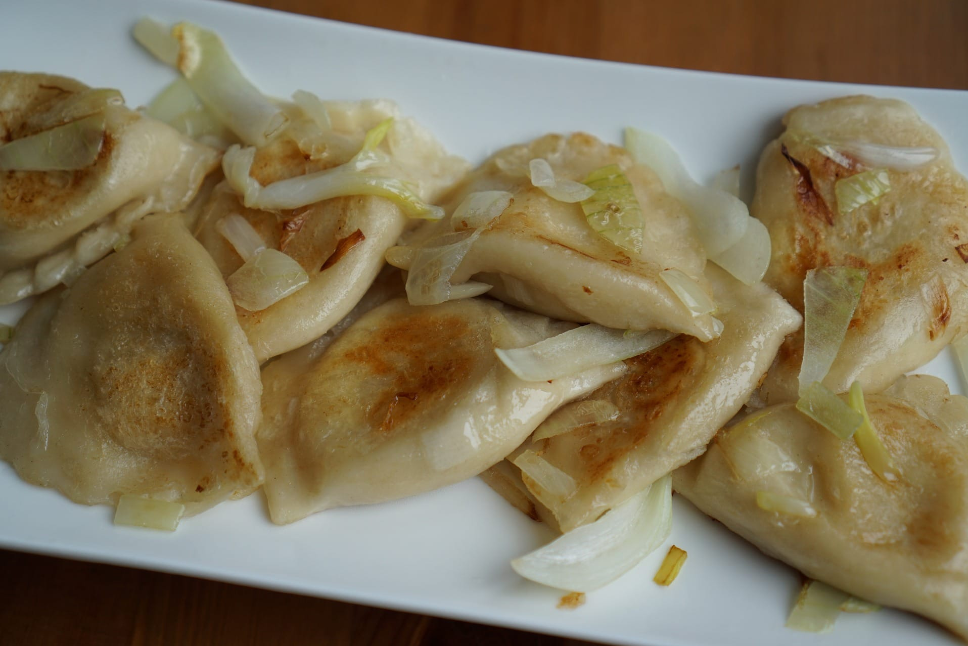 Porcja smażonych pierogów z cebulką na białym talerzu, widoczne delikatne przypieczenia ciasta i błyszcząca cebula.