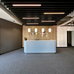 Nowoczesne biuro z recepcją w odcieniach szarości i błękitu, betonowa ściana w tle z logo firmy, sufit z czarnymi belkami i oświetleniem liniowym.