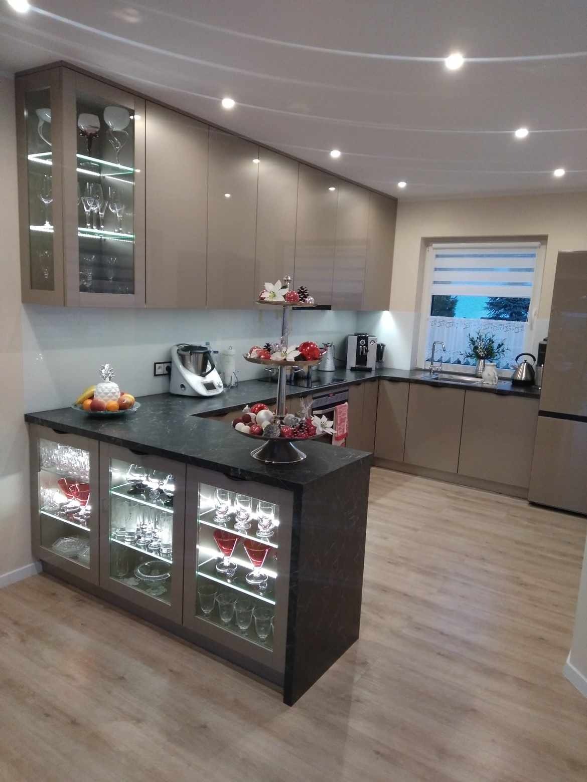 Beżowa kuchnia z barkiem, szklanymi witrynami podświetlonymi LED, czarnym blatem i dekoracjami świątecznymi. Nowoczesne oświetlenie sufitowe.