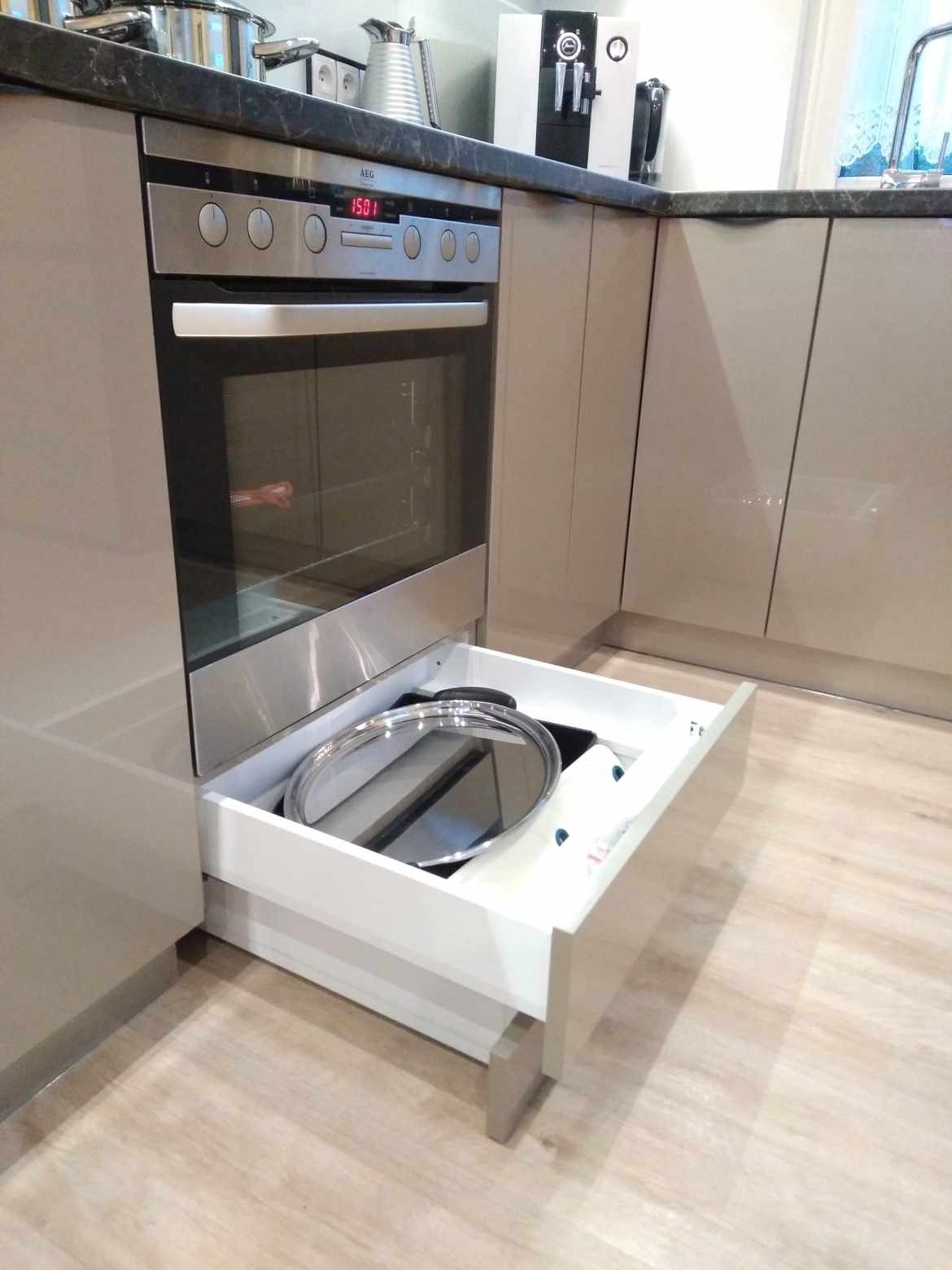 Beżowa zabudowa kuchenna z piekarnikiem AEG i wysuwaną szufladą, w której znajduje się pokrywka i akcesoria kuchenne. Blat z ciemnego kamienia.