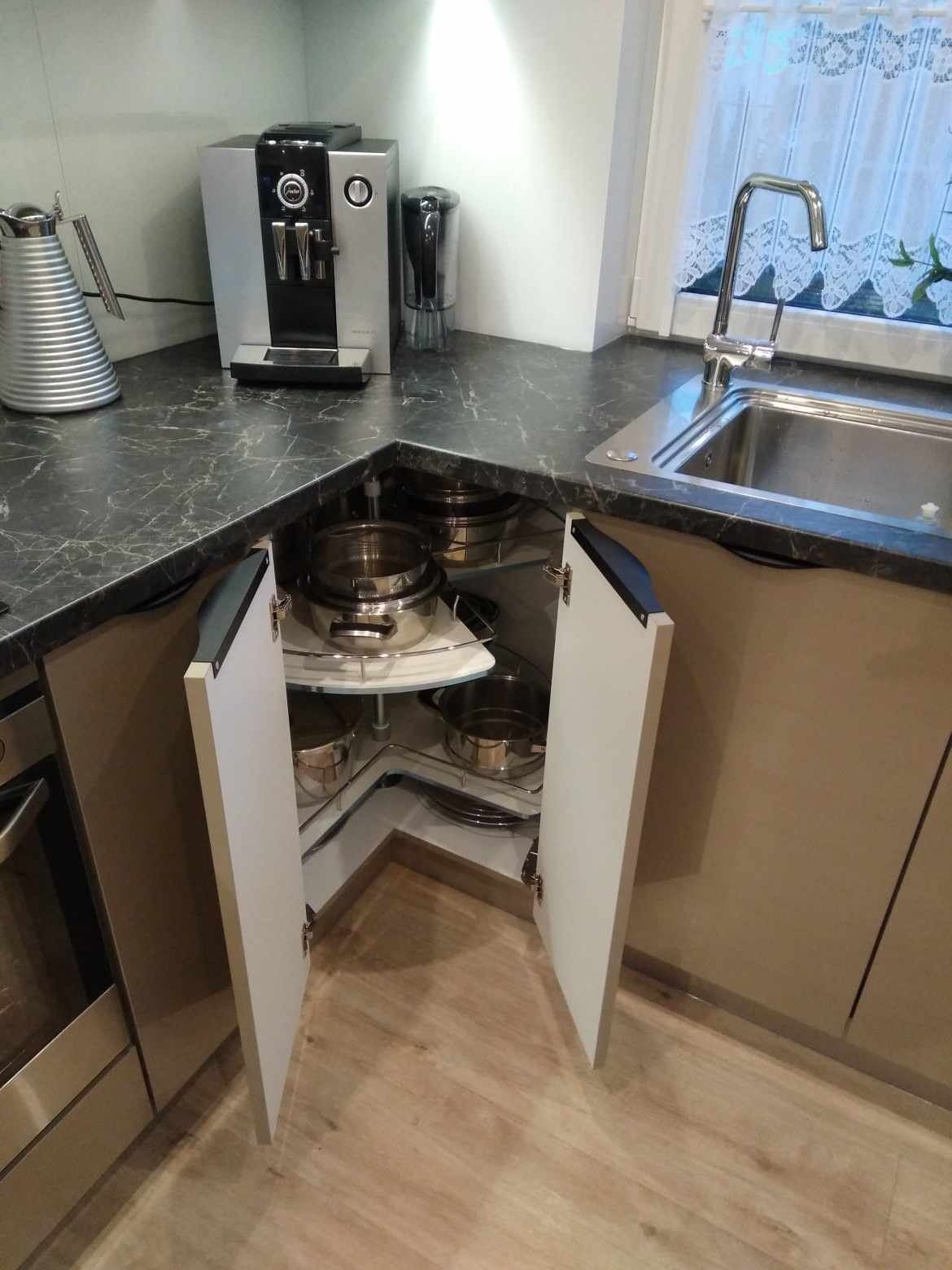 Kuchnia z blatem imitującym marmur, z otwartą szafką narożną z obrotowymi półkami, na których stoją garnki. Widoczny zlew i ekspres do kawy.