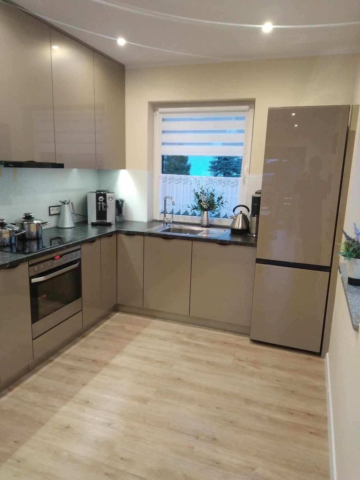 Beżowa kuchnia w kształcie litery L z ciemnym blatem, sprzętem AGD i oknem z roletami. Widok na zlew, płytę grzewczą i piekarnik. Jasna podłoga.