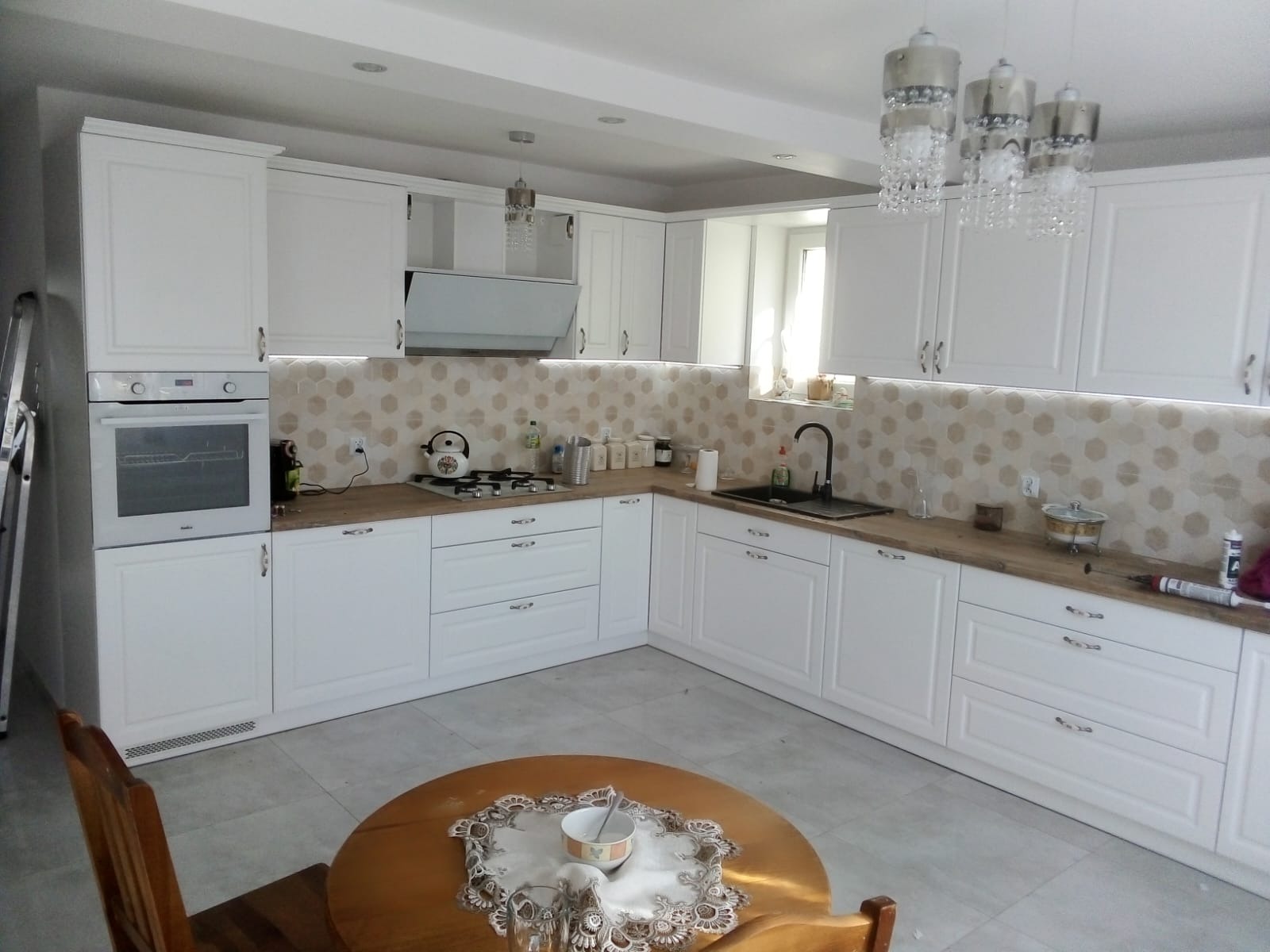 Jasna kuchnia na wymiar z drewnianym blatem i heksagonalnymi płytkami. Widoczne oświetlenie LED podszafkowe i okrągły stół z nakryciem.