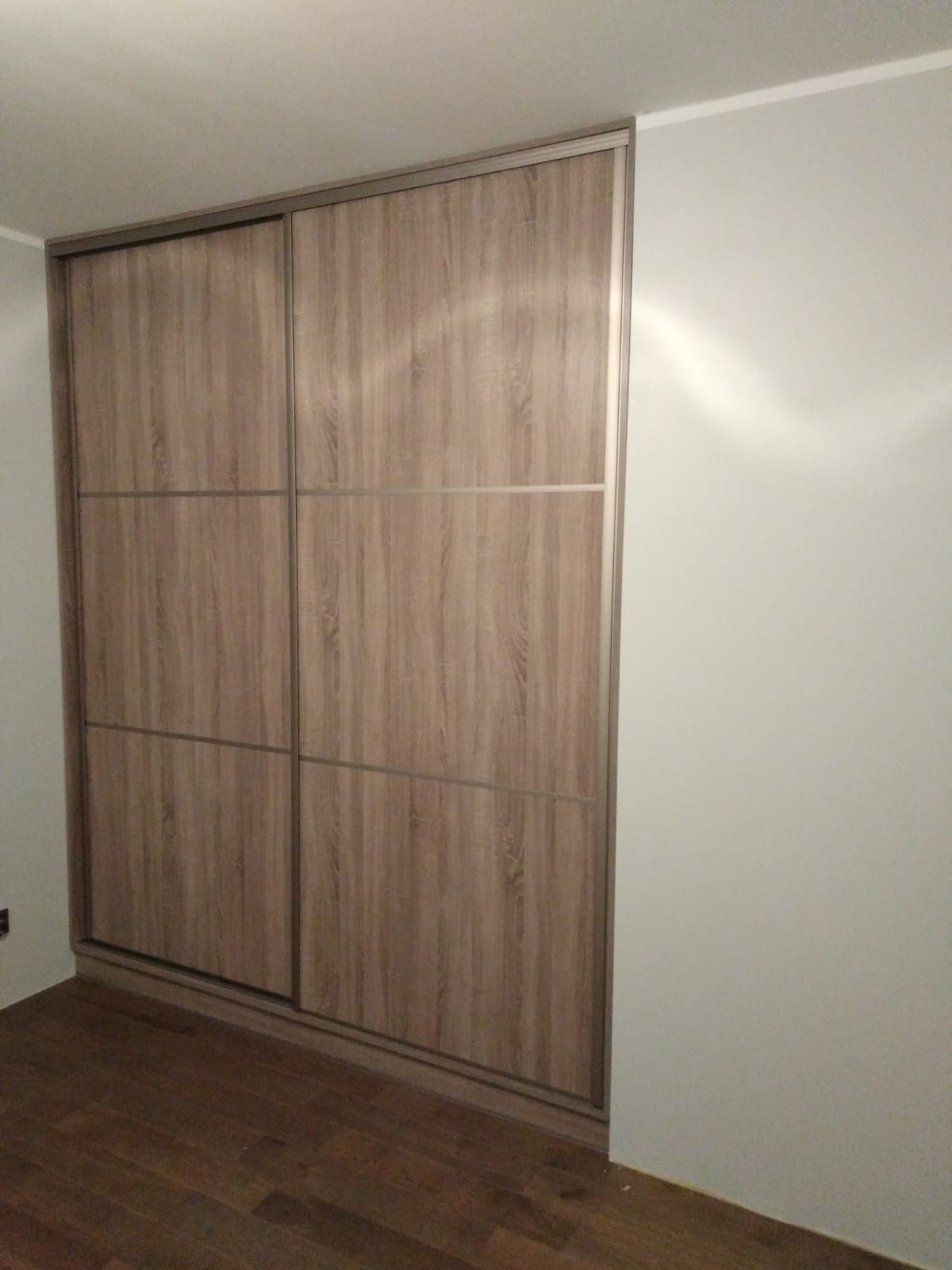 Beżowa szafa wnękowa z przesuwnymi drzwiami z drewnianym wzorem, wkomponowana w jasne wnętrze z parkietem, widoczna z boku, z delikatnym oświetleniem.