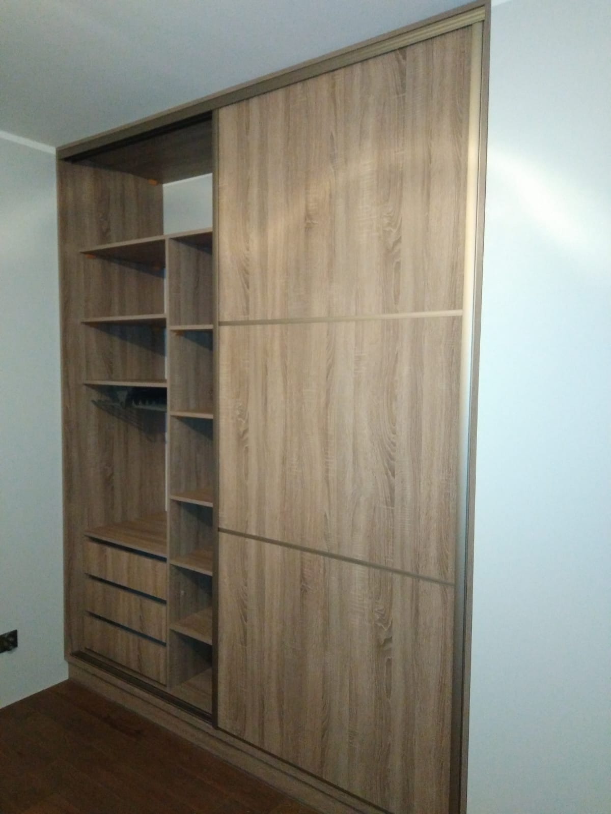 Jasna szafa wnękowa z przesuwnymi drzwiami i otwartą częścią z półkami oraz szufladami, wykończona w odcieniach drewna.