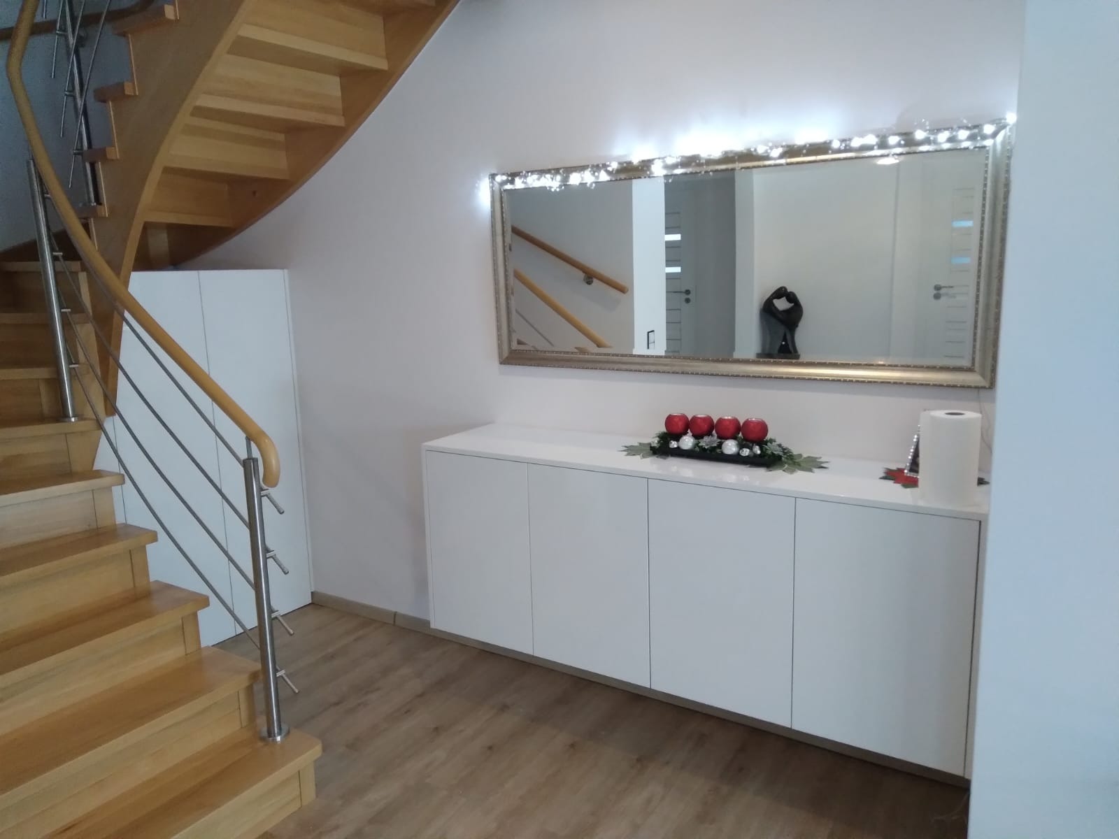 Biała szafka pod schodami z dekoracją świąteczną, lustro w złotej ramie z lampkami, drewniane schody z metalową barierką, parkiet na podłodze.