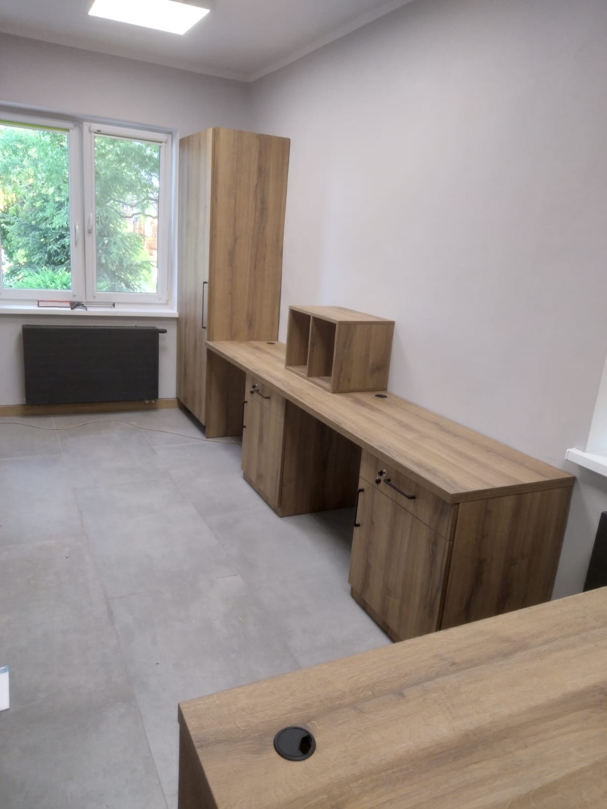 Jasne biuro z szafą i długim blatem roboczym z drewna w odcieniach brązu. Minimalistyczny design, szare płytki podłogowe, okno z widokiem na zieleń.