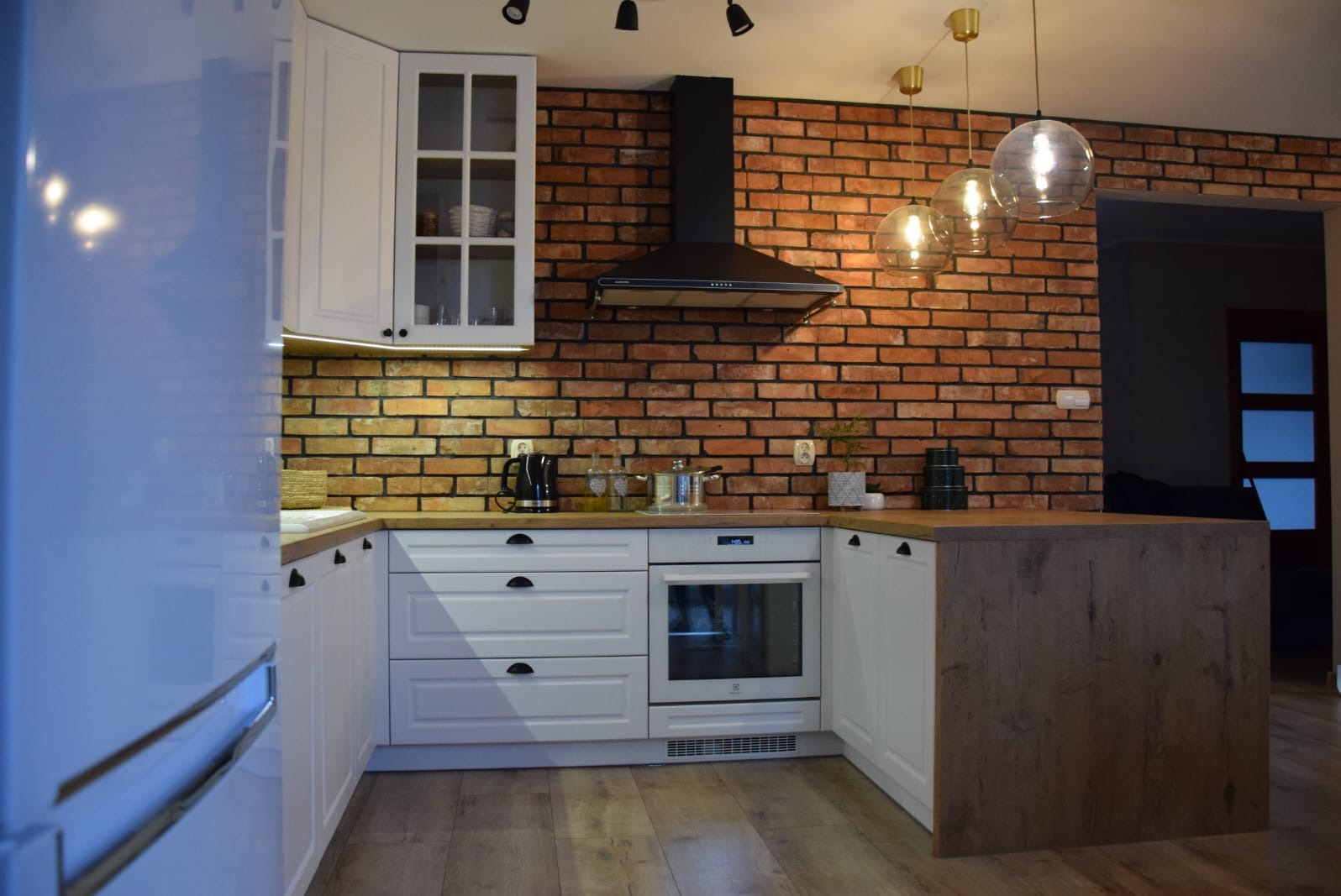 Biała kuchnia z ceglaną ścianą, drewnianym blatem i wyspą. Ciepłe oświetlenie podkreśla rustykalny charakter wnętrza. Widoczne sprzęty AGD i dekoracje.
