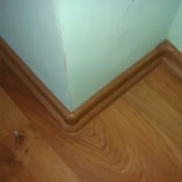 Fragment wykończonej podłogi z drewnianymi panelami i listwą przypodłogową w rogu pomieszczenia, widoczny fragment ściany pomalowanej na jasnozielono.