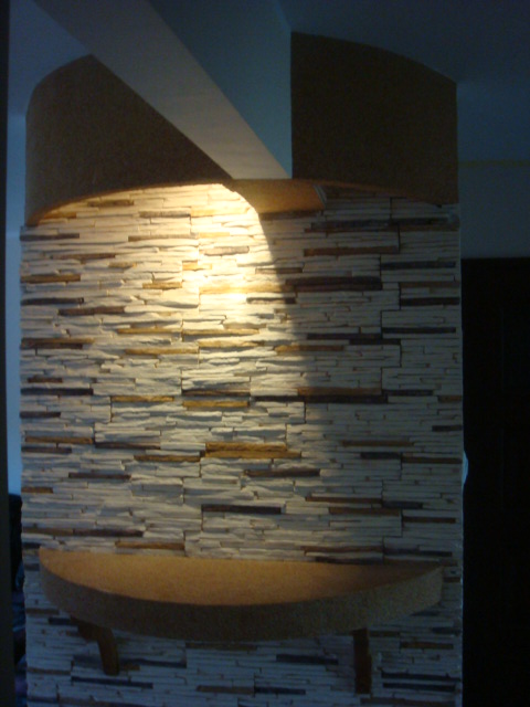 Dekoracyjna ściana wykończona kamieniem elewacyjnym z półkami z naturalnego drewna, oświetlona punktowym światłem.