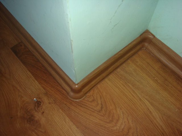 Fragment wykończonej podłogi z drewnianymi panelami i listwą przypodłogową w rogu pomieszczenia, widoczny fragment ściany pomalowanej na jasnozielono.