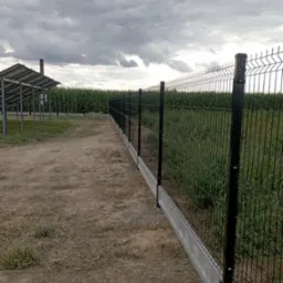 Czarny panelowy płot z podmurówką w Rokicinach, widok z boku na tle pola kukurydzy i pochmurnego nieba.